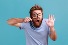 Portrait Of Funny Bearded Man Holding Magnifying Glass And Looking At Camera With Big Zoom Eye, Curious Face, Waving Hand, Saying Hi. Indoor Studio Shot Isolated On Blue Background.