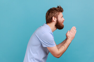 Please, I'm begging. Side view of bearded man holding arms in prayer, asking help or forgiveness with imploring eyes, sincere asking permission. Indoor studio shot isolated on blue background.