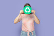 © khosrork - Portrait of unknown anonymous woman wearing striped T-shirt holding green recycling sign and hiding her face behind ecology symbol. Indoor studio shot isolated on purple background.