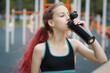 © upparadox - Young sporty slim girl in black sport top drinking water from bottle on a sportsground during training or jogging, urban health and wellness  lifestyle close up portrait
