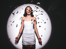 Young Beautiful Smiling Brunette Female In Trendy Evening Silver Dress. Sexy Carefree Woman Posing Near White Wall In Studio In A Circle Of Light.Fashionable Model With Bright Makeup. Falling Confetti