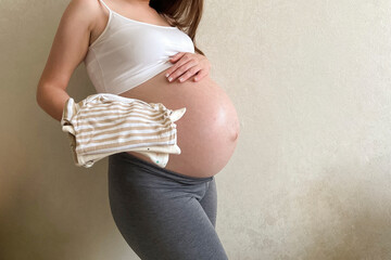 A beautiful pregnant girl is holding a stack of clothes for a newborn. Preparation for the appearance of the baby. Children's shopping. Copy space