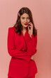 © Look! - Beautiful young caucasian brunette girl looks into camera holding hand to face on pink background. Model wears red jacket to meet her partner. Lifestyle, feminine beauty concept