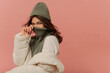 © Look! - Cheerful young caucasian girl looking into cameras covers her face with collar of sweater on pink background. Brunette with wavy hair wears grey hat, sweater and white jacket.