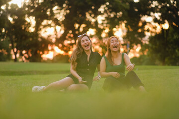 Two young Asian women, both with long wavy hair, howl with laughter. They are sitting beside each other on the grass on a beautiful summer day at sunset.