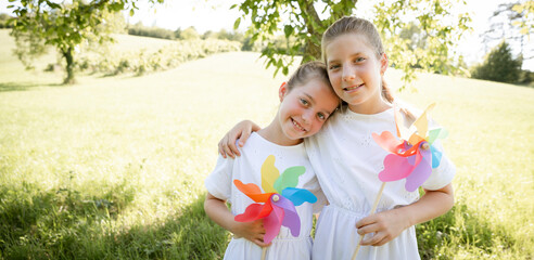 two pretty girls, sisters with white dresses holding colorful pinwheels and standing in green meadow in front of a walnut tree and are happy