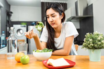 Wall Mural - Unhappy female looking to organic greens vegetable salad in weight loss diet and wellness on table. Tired woman dislike eat bored with food healthy salad breakfast in kitchen. Diet food concept.