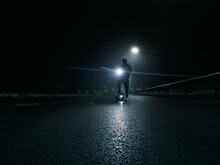 A Man Rider On An Electric Scooter Is Standing On The Road, A Foggy Autumn Night, The Light From The Lanterns