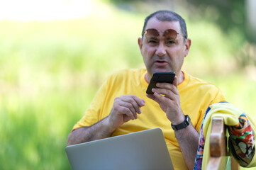 Image of content european man 50s in casual clothing sitting on bench in park and talking on cell phone while using silver laptop