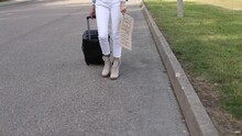 Slim Legs Of Woman With Suitcase And Cardboard Poster Walk Along Empty Road Closer To Camera While Wait Passing Car. Lady Escape From City To Go Anywhere. Travelling, Freedom, Hitchhiking, Vacations