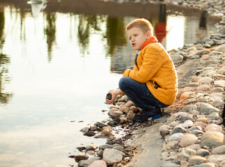 Portrait of red-haired teenager boy in yellow clothes near lake in city park at sunset. Child playing with pebbles