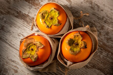 Three Ripe Persimmons Wrapped In A Sack Of Esparto Grass On Rustic Wood.