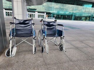 Two wheelchairs are lined up in front of a hospital or shopping mall waiting to serve the elderly, sick or handicapped. The wheelchair is made of steel with dark blue seats and large black wheels.
