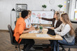 © sofiko14 - Group of four multicultural female economists looking on big monitor with various graphs during working meeting at office. Businesswomen partners using modern technology during negotiation.