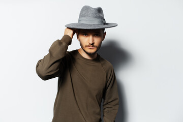 Wall Mural - Studio portrait of young man holding wireless earbuds and charger case in hands on white background, wearing sweater and grey hat.