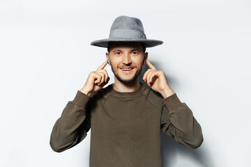 Studio portrait of young happiness man, listening the music via wireless earbuds on white background, wearing sweater and grey hat. Touching earphones with fingers.