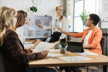 Caucasian blond woman in white shirt and eyeglasses showing financial statistic of enterprise during working meeting with diverse female colleagues. Cooperation, technology and people concept.