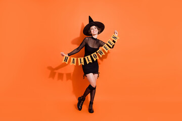 Wall Mural - Full length photo of adorable young lady hold garland flags wear stylish black halloween witch clothes isolated on orange color background
