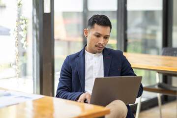 Wall Mural - Asian Business man Using laptop computer, Successful Asian entrepreneur smiling in satisfaction as he checks information on his laptop computer while working