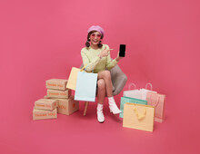 Happy Asian Teen Woman Sitting On Sofa Holding Shopping Bags And Smartphone Isolated On Pink Background, Shopper Or Shopaholic Concept.