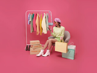 Happy Asian teen woman sitting on sofa holding shopping bags and smartphone isolated on pink background, Shopper or shopaholic concept.