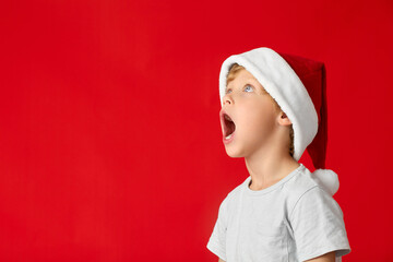 Boy opened his mouth wide in surprise, looking up. Child in Christmas hat looks up in surprise against the background of red wall