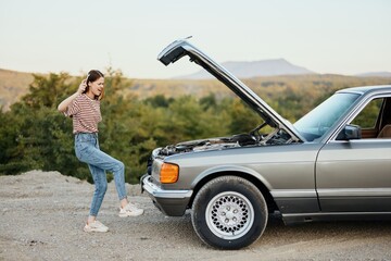 A woman opens the hood of a broken down car and tries to find the cause of the breakdown on a road trip alone and kicks the car in anger