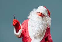 Santa Claus Blue Studio Background In Red Cap With A White Beard Looks At The Camera And Points To Side With Pointer. Copy Space. Pointing To The Left, Leaning To The Right.
