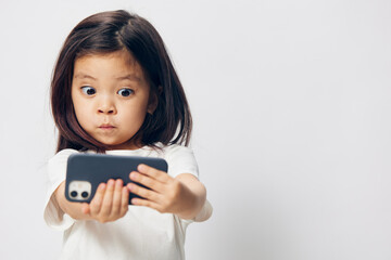 Wall Mural - cute, funny, little girl in a white t-shirt stands on a light background with a smartphone in her hand and takes a selfie. Horizontal studio photography with blank space for advertising mockup insert