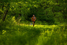 Woman Running In A Green Field In The Forest With A Dog