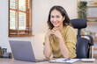 © David - Portrait of smiling asian woman working at home on some project, her is sitting at a table looking at camera, writing ideas with her laptop, freelancer indian businesswoman work on documents.