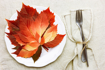 Autumn table setting, linen napkin, plate, knife, fork on  tablecloth. Top view white plate decorated fall red leaves and cutlery.  Autumn home and table decor. Seasonal holiday natural decor