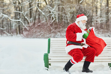 Wall Mural - Side view portrait of traditional santa Claus holding bag with presents while sitting on bench outdoors, copy space