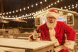 Portrait of traditional Santa Claus looking at camera with hot coffee cup in park with fairy lights in background, copy space