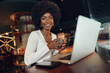 © fotofabrika - Smiling young african woman sitting with laptop in coffee shop