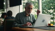 © Marco - Older executive opening laptop seated at cafe store working remotely. Middle aged senior man browsing internet online with computer at coffee shop