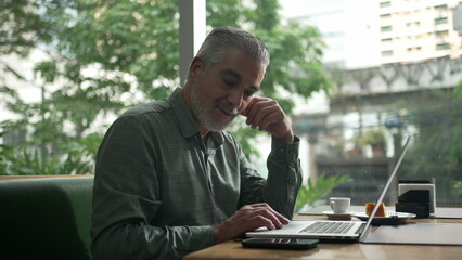 Wall Mural - Middle aged man working at coffee shop in front of laptop. Older person receiving message notification on phone. Executive person typing on computer