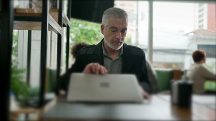 Wall Mural - Male entrepreneur opening laptop at coffee shop table. Older middle aged person working remotely at cafe place using modern technology computer