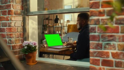 Aufkleber - African Man Working on a Laptop Computer with Green Screen Mock Up Display while Sitting at a Table in Stylish Living Room. Business Manager Working Remotely from Home. View from Outside the Window.