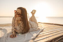 Summer Lifestyle, Young Caucasian Woman Lies On Seashore In Sunglasses On Mat. Pretty Woman In Sunglasses Looks Into Distance With Smile On Her Face. Rest And Recovery Concept.