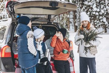 Wall Mural - Four friends teenagers boys and girl with thermos sitting in trunk of car decorated for Christmas and New year. Winter picnic in snowy forest. Road trip and local travel. Friends hanging out together