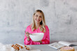 © VikaNorm - Smiling woman whisking eggs with a whisk in a white mixing bowl on a grey background. A young blonde woman in a pink blouse prepares pastries in the kitchen according to the recipe in the book.