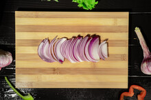 Purple Onion, Thinly Sliced On A Striped Wooden Board. Top View Of Slices Of Juicy Onion, Lettuce, Garlic, Pieces Of Red And Green Sweet Pepper. Sliced Wet Vegetables On A Black Table Close Up.