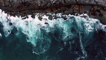 Aerial Top View Of The Beautiful Ocean Rocky Shore With Rolling Waves. Drone Is Hovering Over Rolling Ocean Waves To The Rocky Shore. Drone Hovers Over Surface Of The Water Along The Rocky Shore.