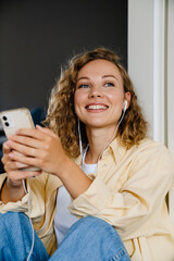 Wall Mural - White woman using cellphone with earphones sitting on floor at home