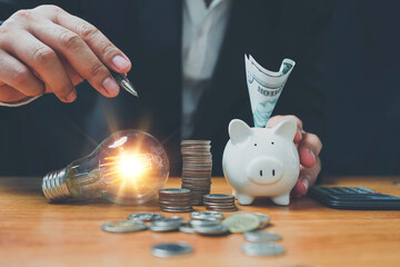 saving energy and money concept. idea for save or investment.businessman holding a pen pointing a light bulb to saving money wealth saving money coins on table concept financial.