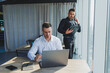 © DSMT - Two male focused colleagues discussing business project looking at laptop screen during meeting in modern meeting room with glass walls