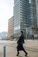 Wall Mural - Girl on the street in a black coat. Stylish girl in a black raincoat on the background of a high glass house