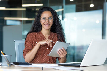 Wall Mural - Happy and successful hispanic woman working inside modern office building, business woman using tablet computer smiling and looking at camera worker doing paperwork.