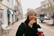 Funny adorable girl in knitted cap and black jacket winking at camera and bitting cookie and drinking coffee while walking around the city in sunny day.
